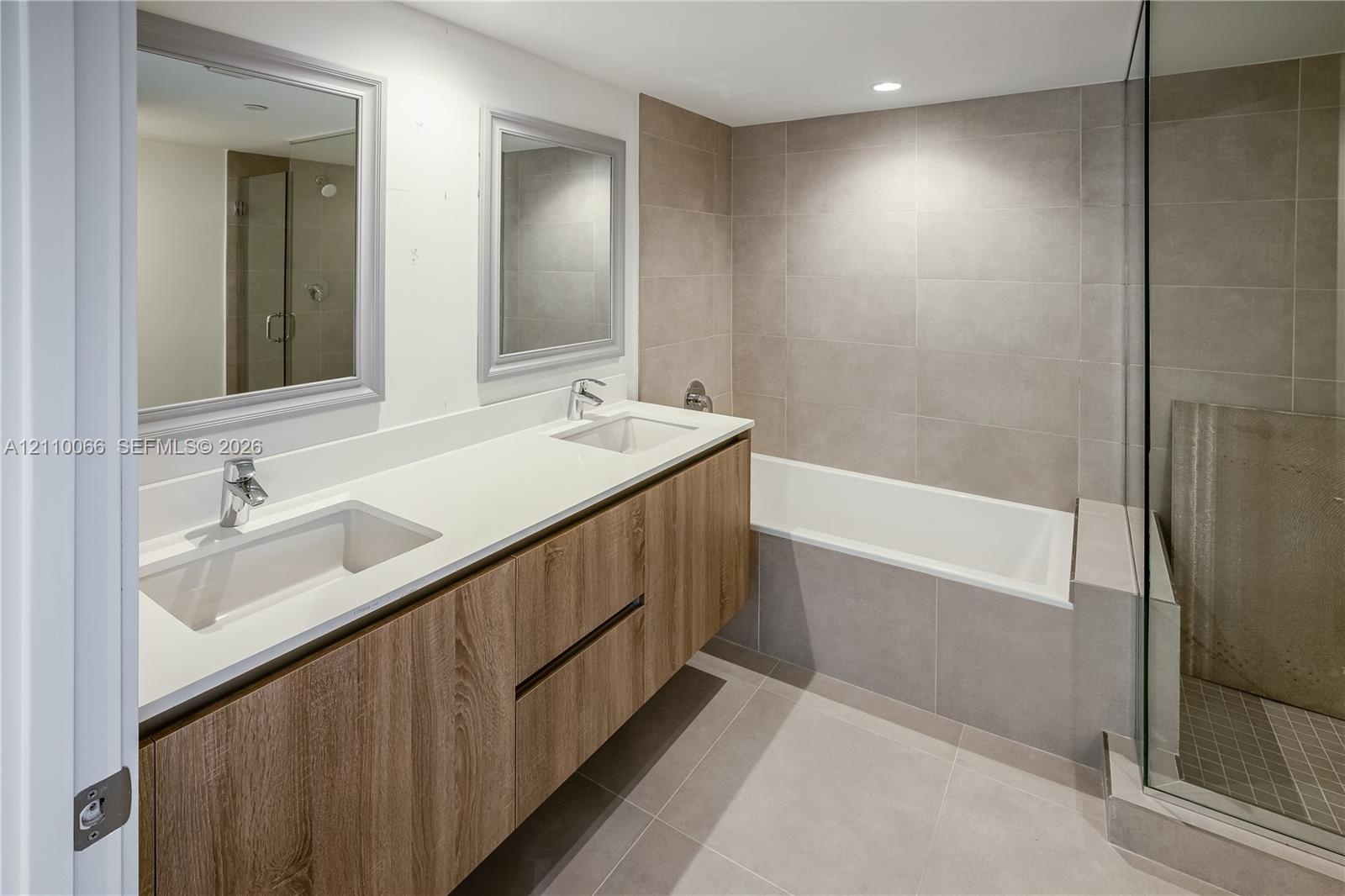 Floating dual-sink vanity with wood-finish cabinetry, solid-surface countertop, separate glass-enclosed shower, and deep soaking tub with neutral tile