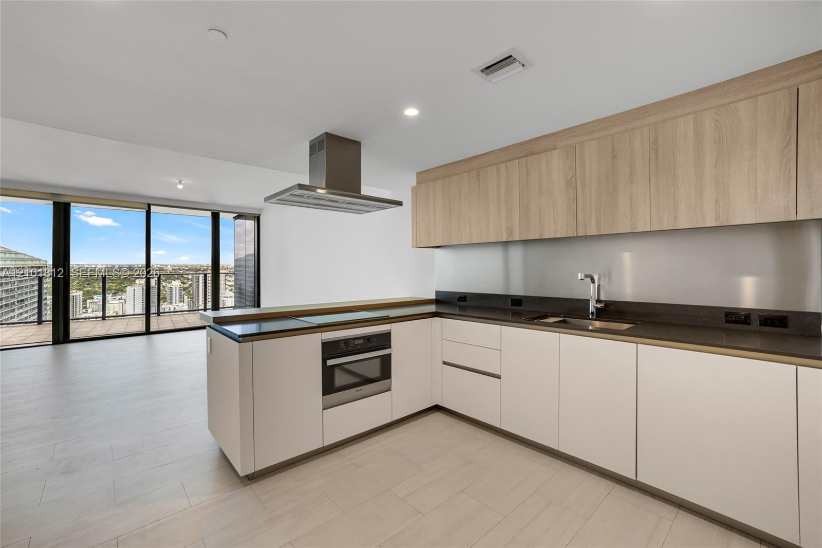 Kitchen featuring flat-panel cabinetry with wood-finish uppers, dark solid-surface countertops, stainless steel wall-mounted range hood, built-in oven
