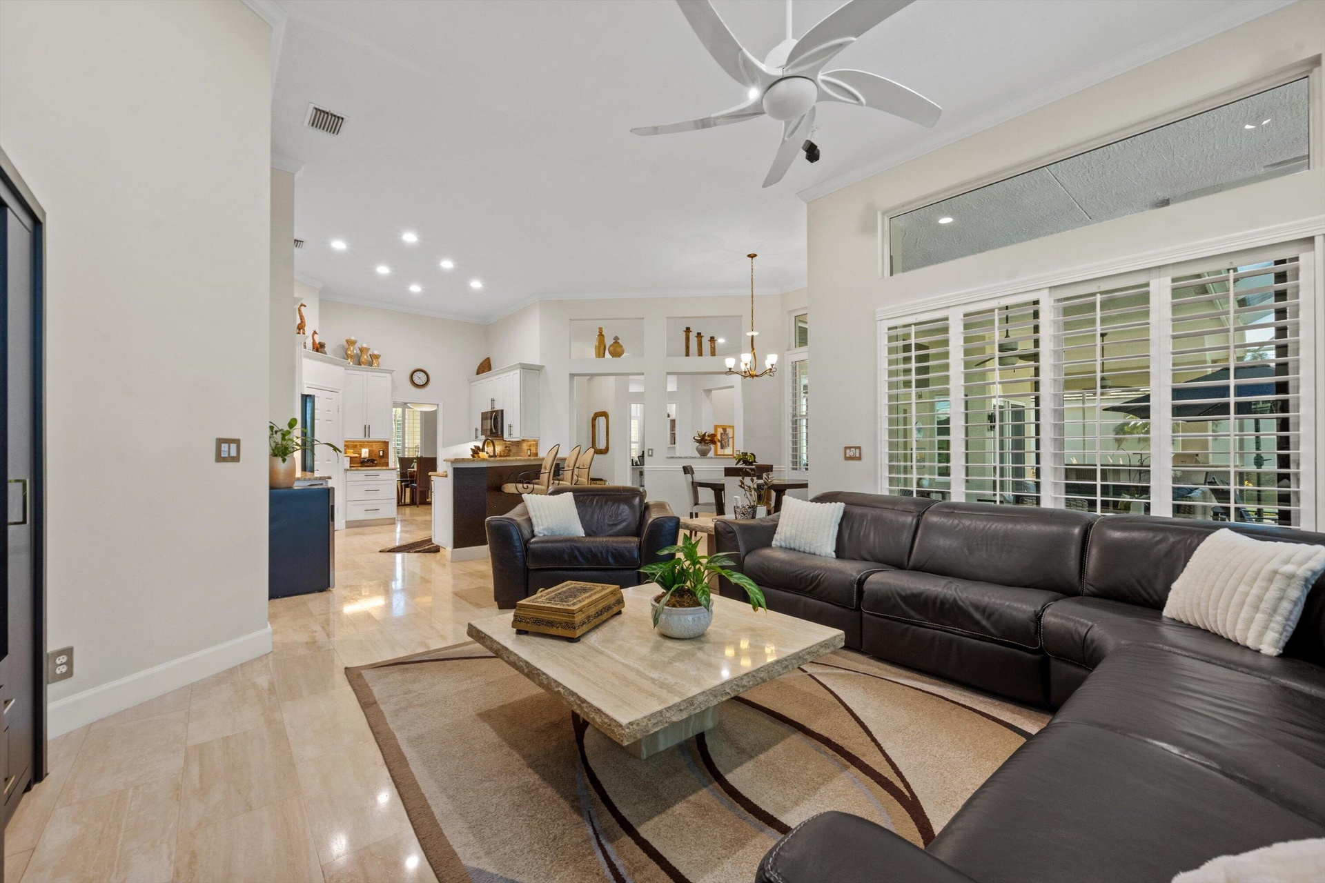 Open-concept interior featuring high ceilings, stone-finish tile flooring, recessed lighting, a multi-blade ceiling fan, and large windows with planta