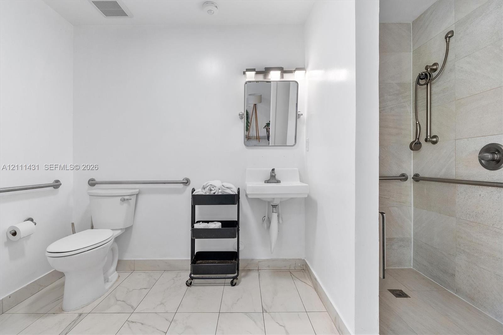 Bathroom featuring light-toned tile flooring, wall-mounted sink, walk-in shower with stone-finish wall tile, and metallic wall-mounted grab bars