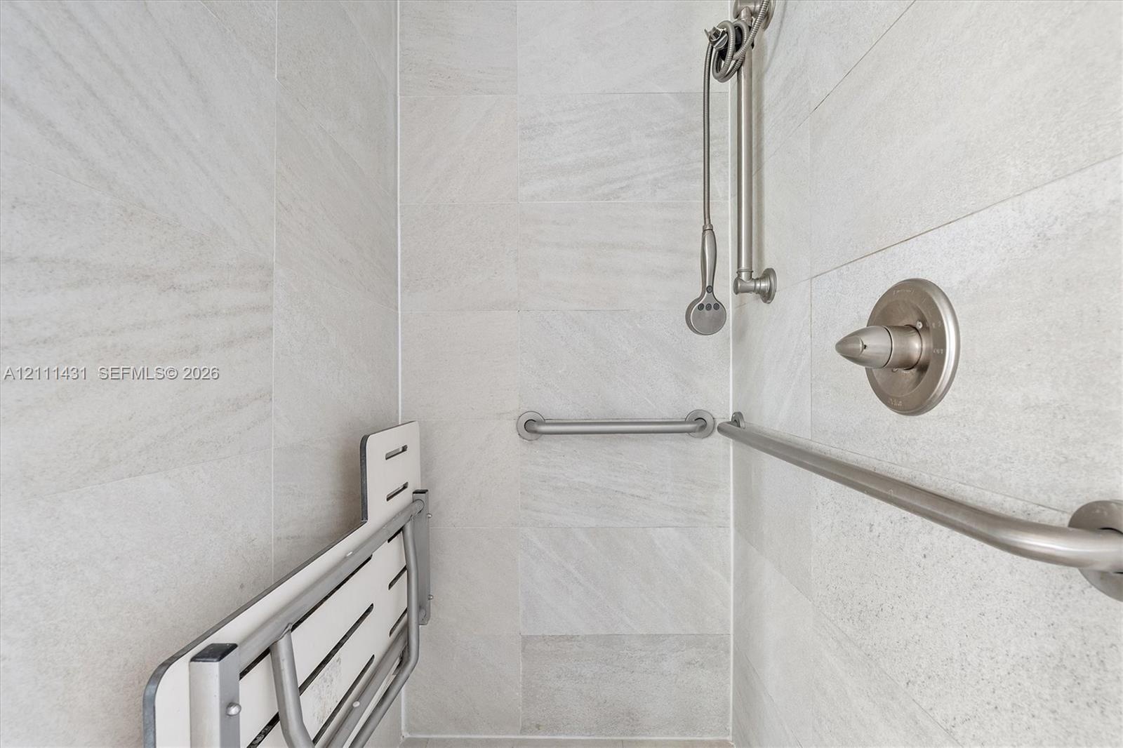 Shower enclosure featuring light grey stone-finish wall tiling, brushed nickel hardware, a wall-mounted shower head on a slide bar, and a fold-down wa
