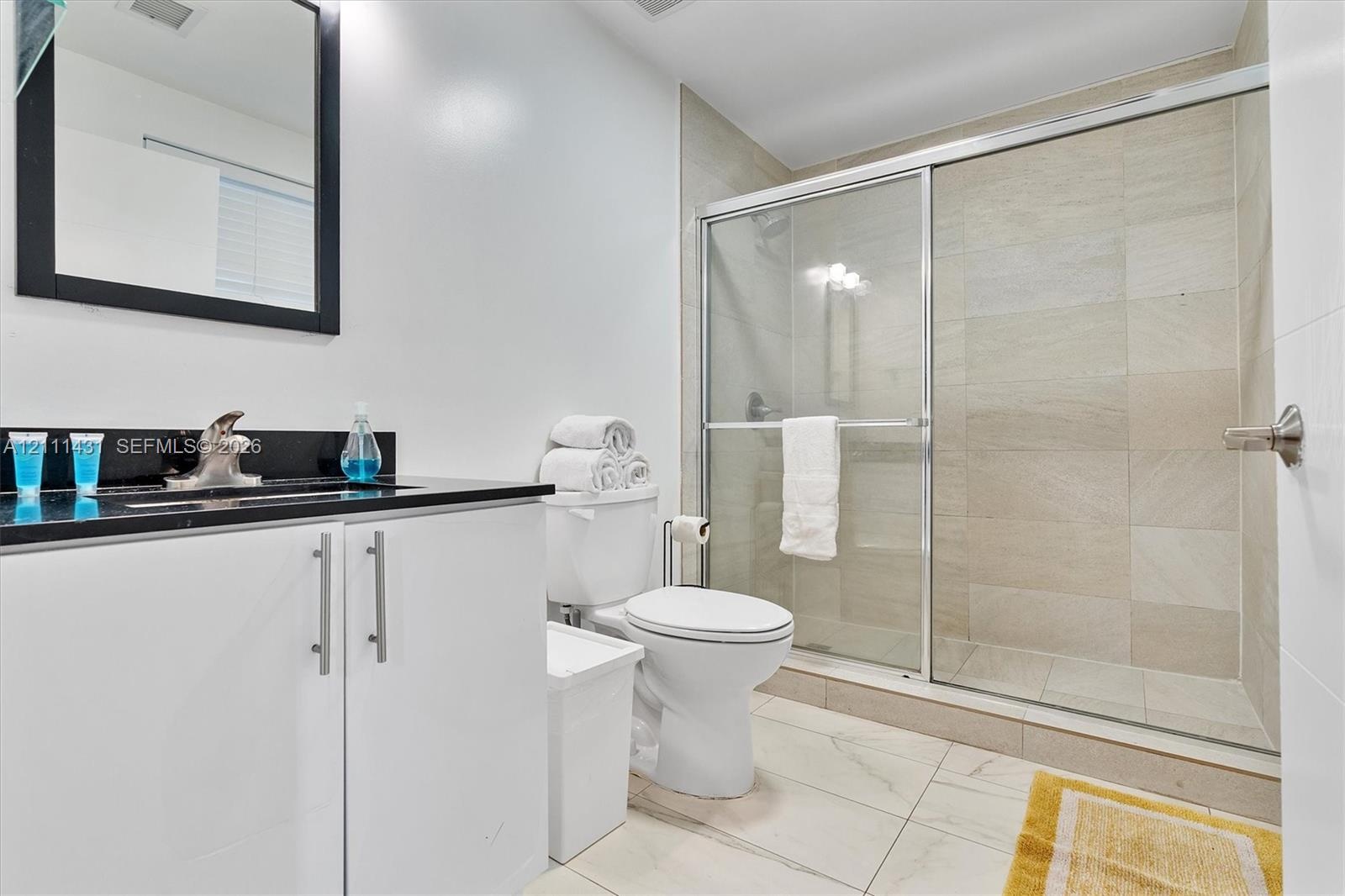 Bathroom featuring a vanity with dark stone countertop, rectangular undermount sink, walk-in shower with sliding glass doors, neutral tile wall surrou