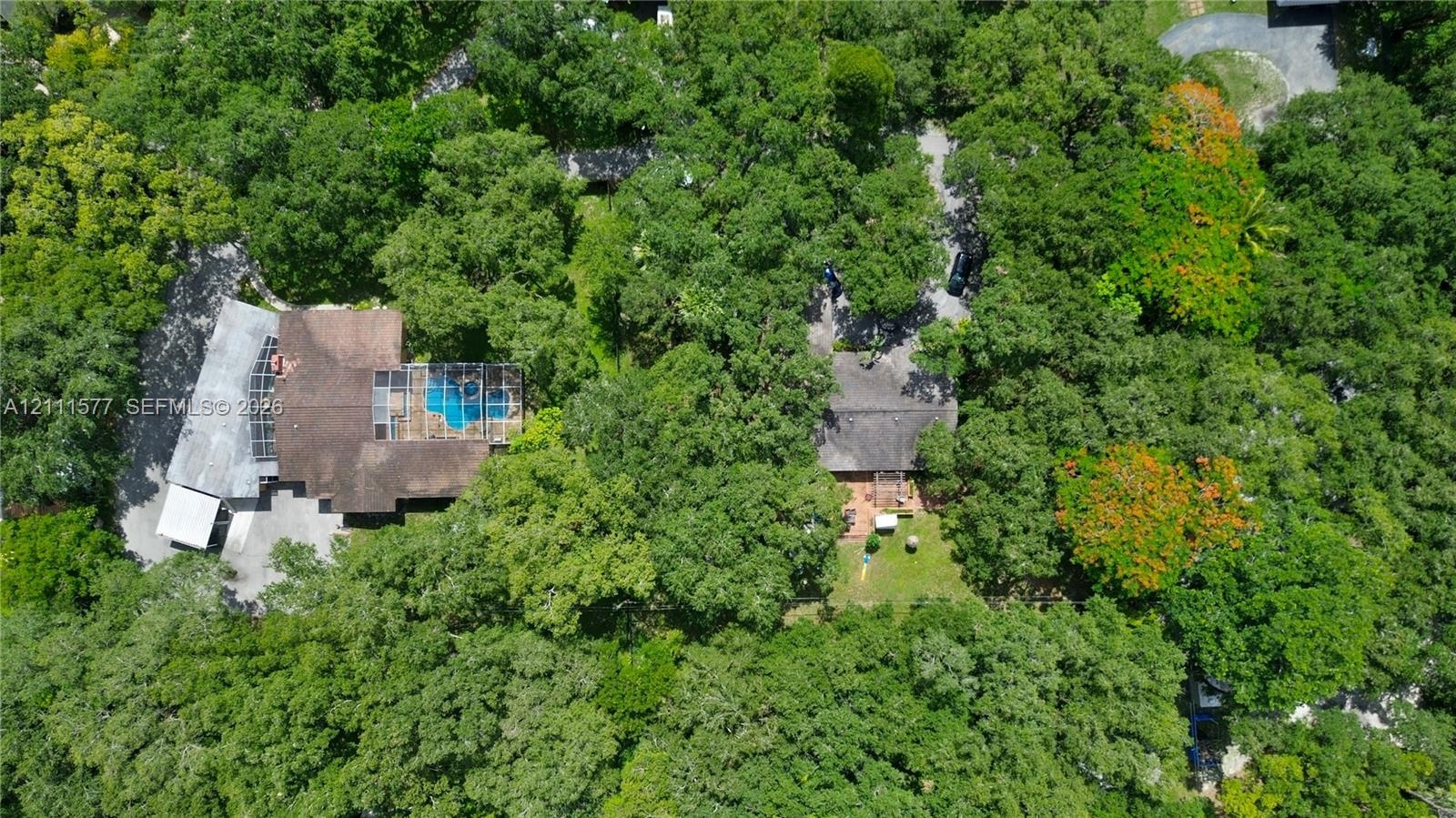 Aerial perspective of a multi-structure property featuring a screened-in swimming pool, shingled roofing, mature tree canopy, and paved driveway