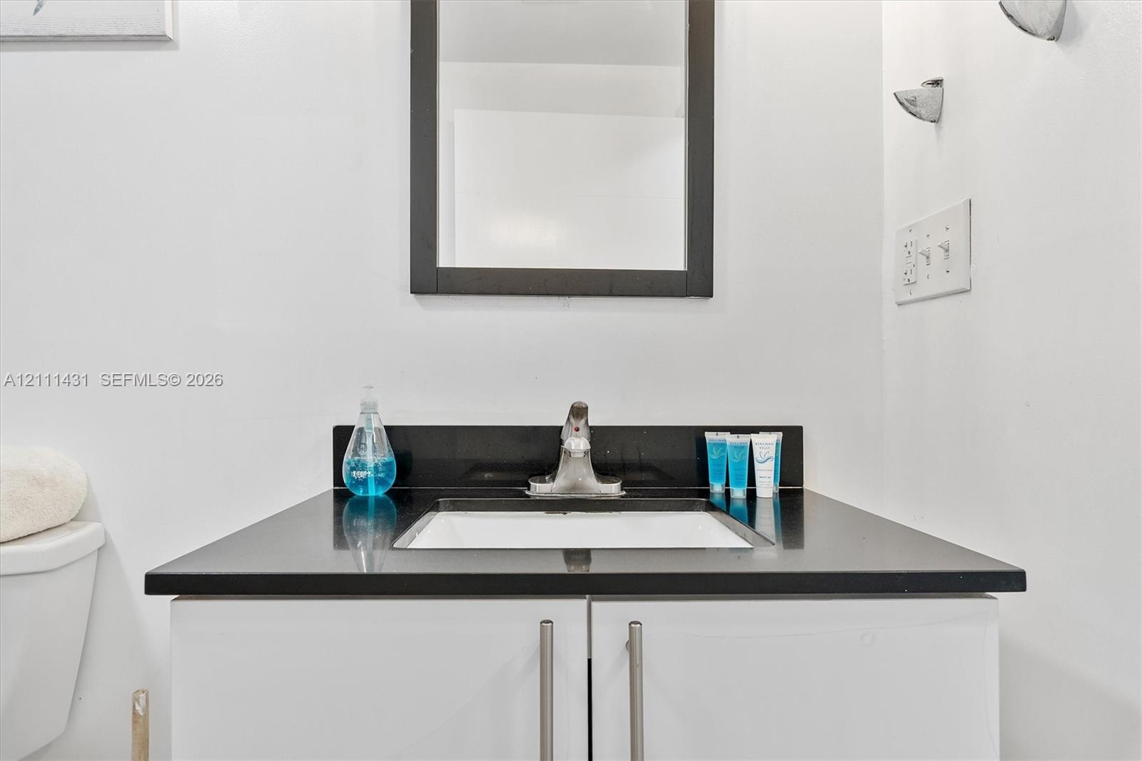 Vanity featuring a dark stone countertop, integrated rectangular sink, polished metal faucet, and white slab-style cabinetry with vertical pulls, topp