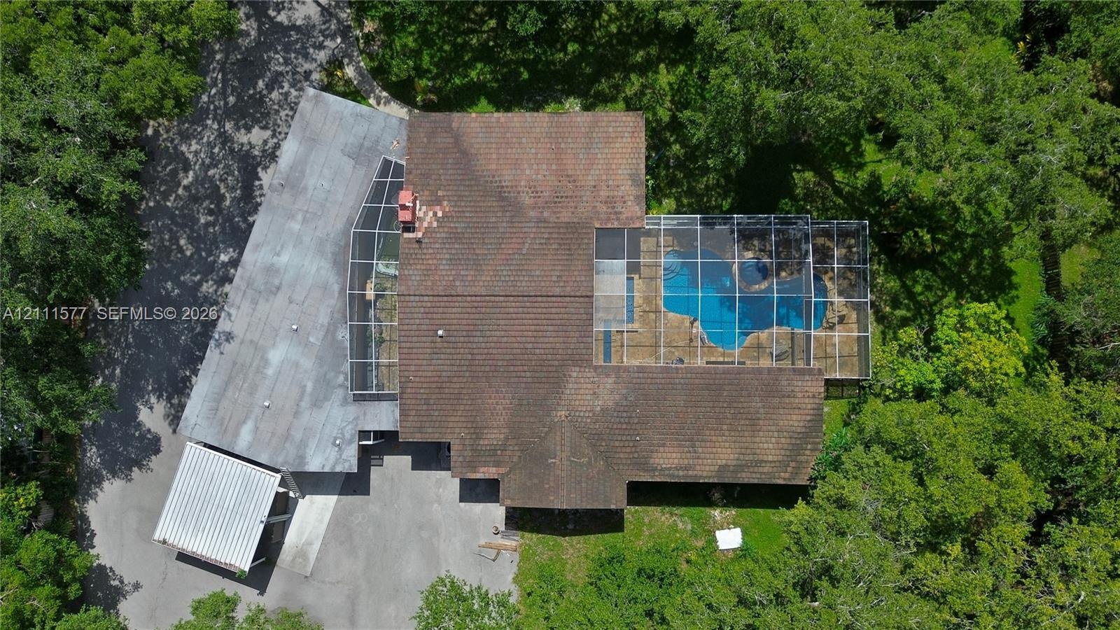 Aerial perspective featuring a multi-level roofline, screened-in swimming pool enclosure, detached covered parking structure, and mature trees on a wo