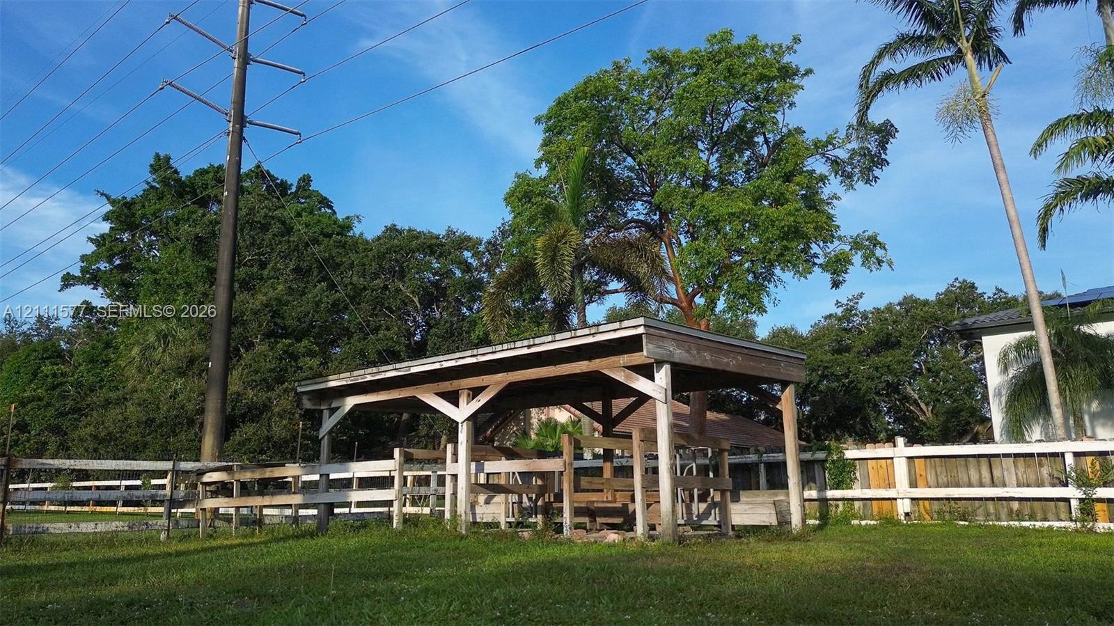 Open-air wood structure featuring a post-and-beam frame, set within a fenced area with mature trees