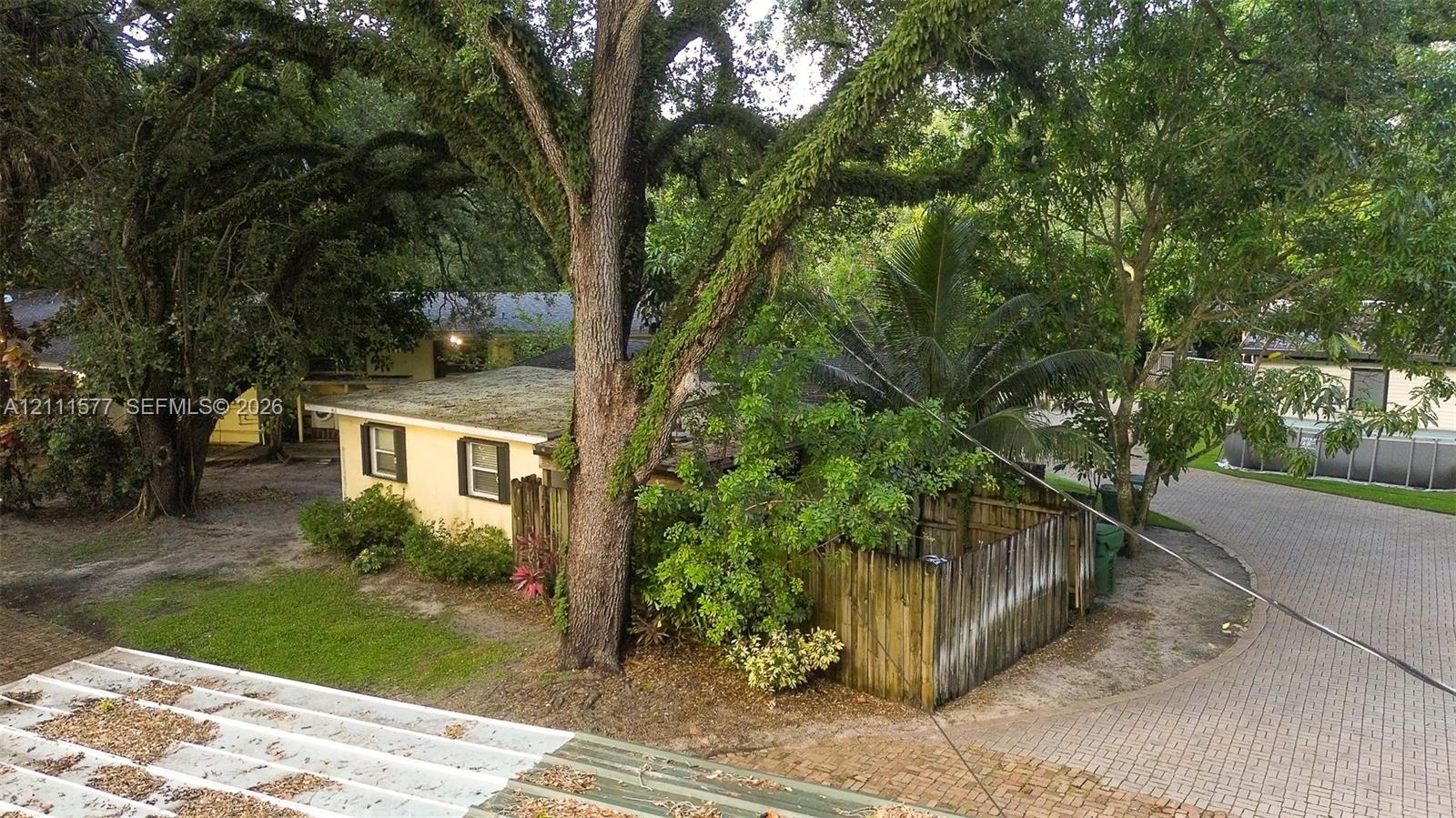 Exterior view featuring a yellow stucco residence, mature oak trees, paved driveway, and perimeter wood fencing
