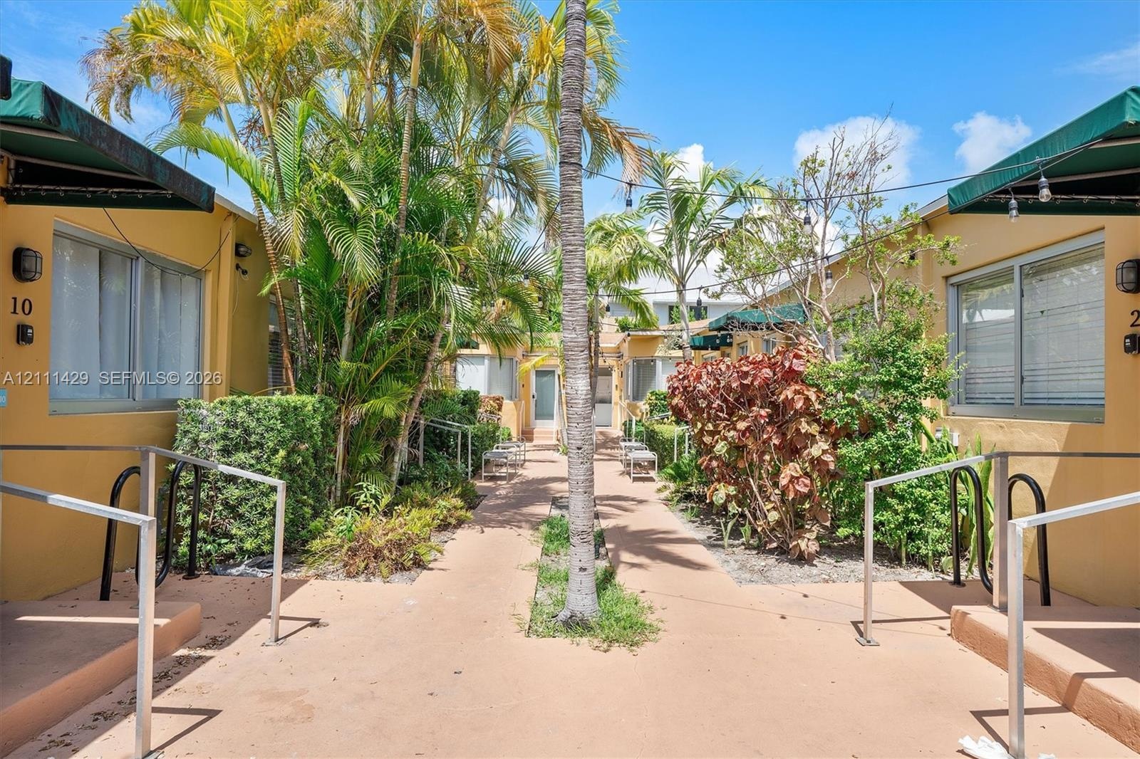 Courtyard featuring paved walkways, mature palm trees, tiered shrubbery, and dark green fabric awnings