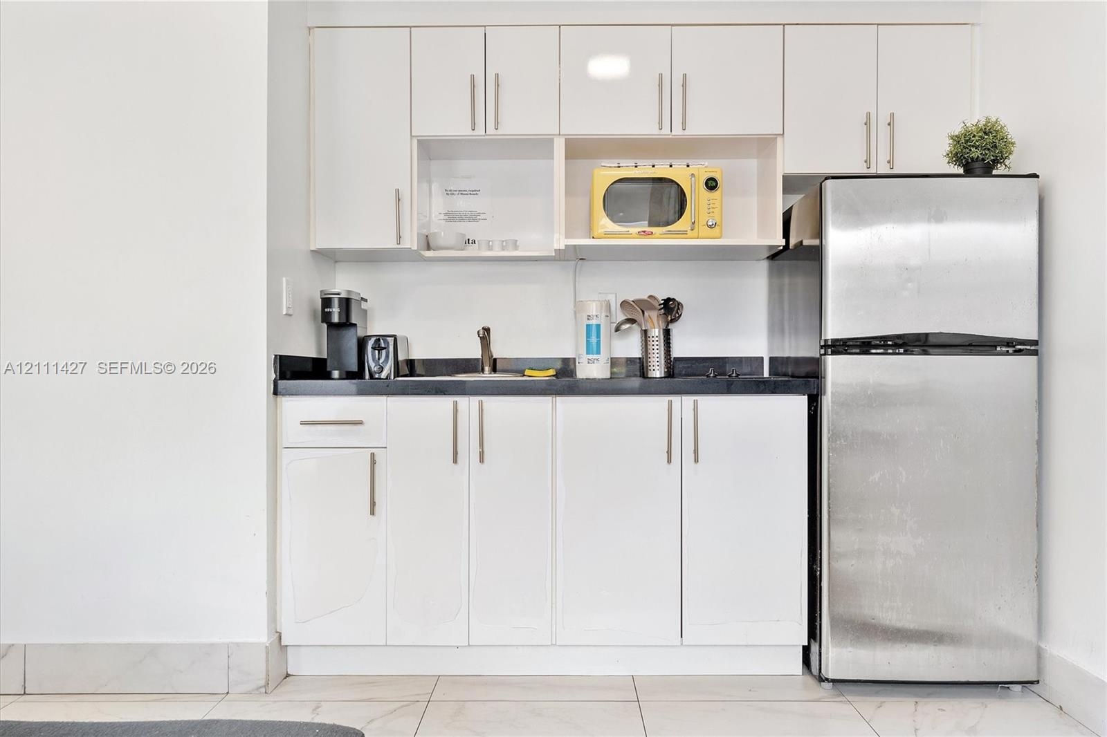 Kitchenette featuring white slab-panel cabinetry, dark stone-finish countertops, stainless steel refrigerator, and ceramic tile flooring