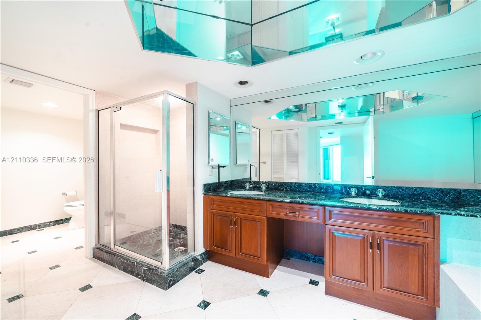 Double vanity with stained wood cabinetry and stone countertops, glass-enclosed shower, expansive wall-to-wall mirror with faceted mirrored accents, a