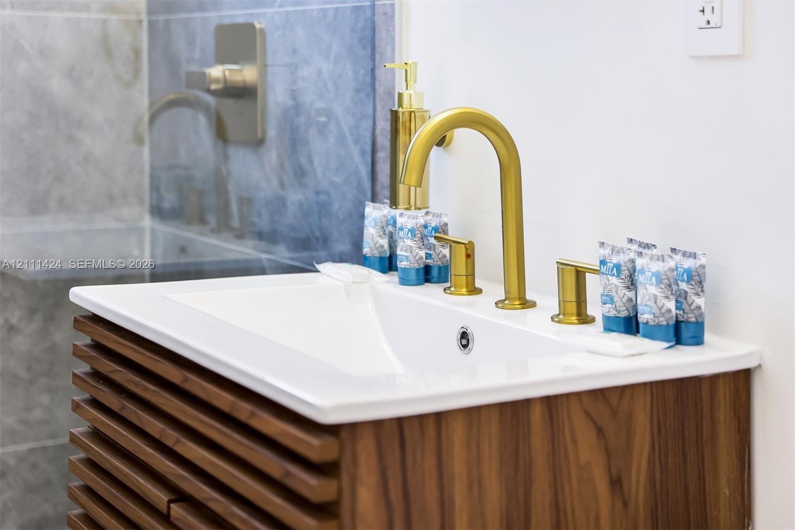 Slatted wood-finish vanity with an integrated white sink, topped by a widespread gold-tone faucet and matching hardware, set against blue-toned stone-