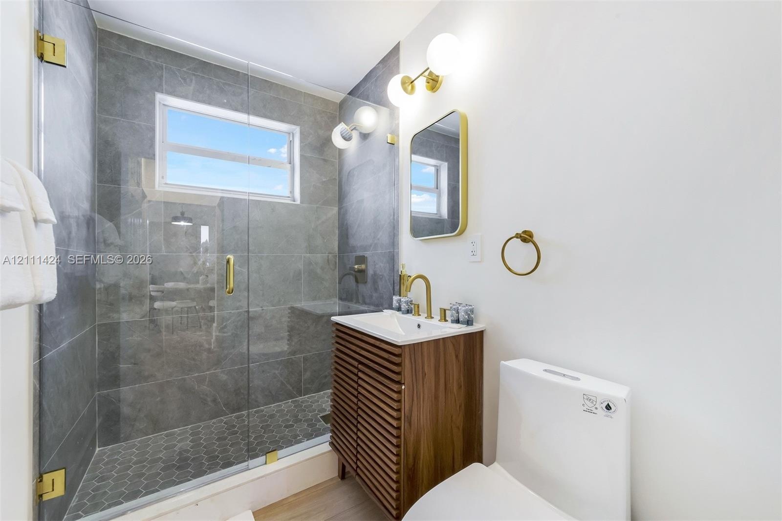 Walk-in shower featuring gray tile surround, glass enclosure, and hexagonal floor tile, complemented by a slatted wood-finish vanity, brass-toned fixt