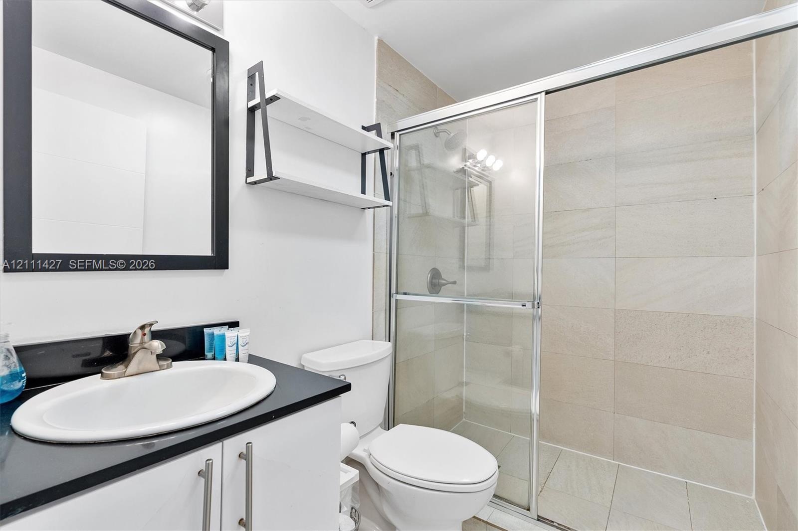 Bathroom featuring a vanity with a dark countertop and integrated sink, a framed mirror, a wall-mounted shelf, and a walk-in shower with a glass door 