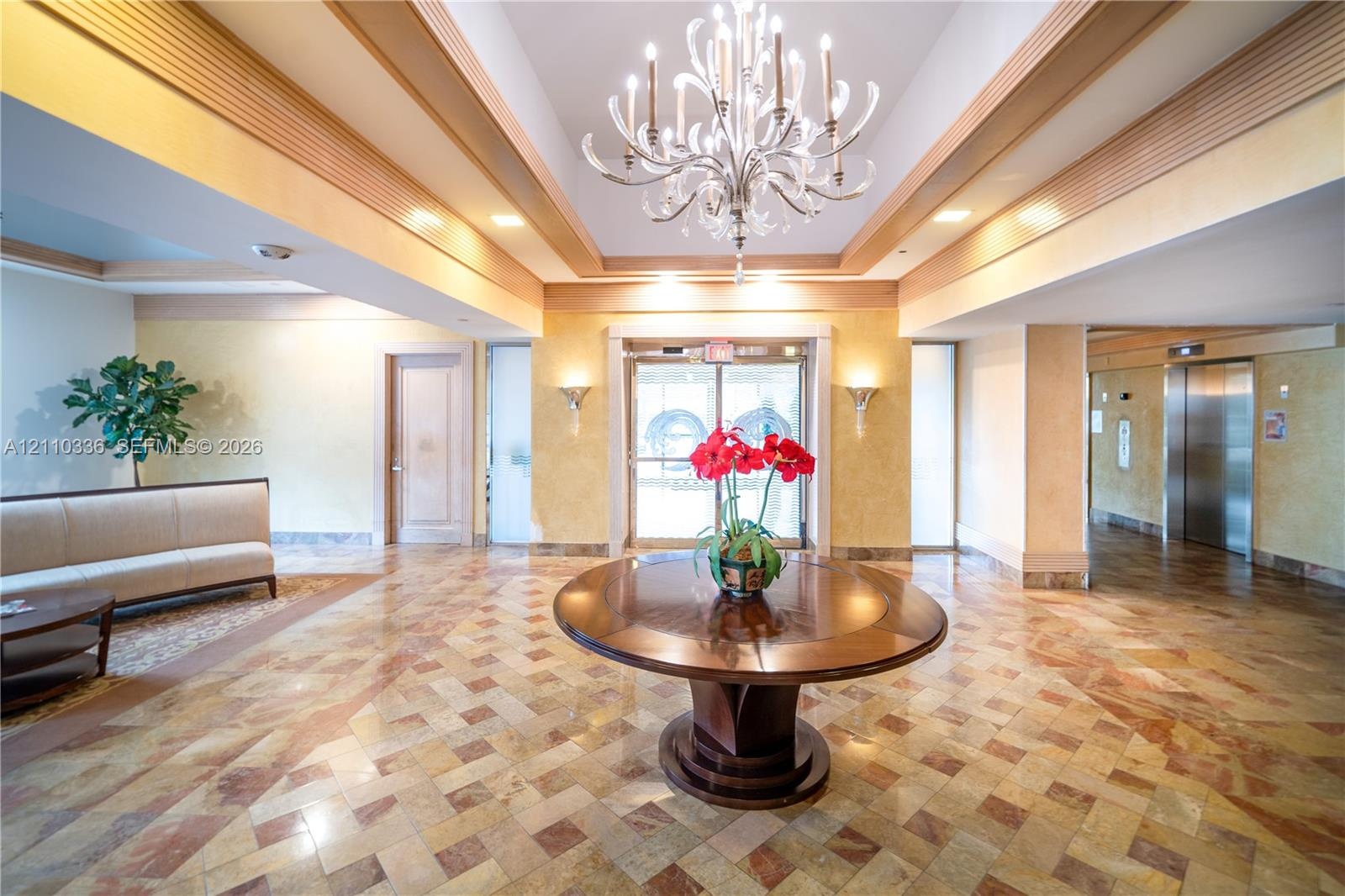 Grand foyer featuring a coffered ceiling with a multi-tiered crystal chandelier, intricate stone tile flooring, and integrated wall sconces