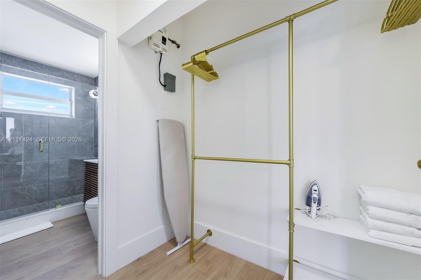 Storage area featuring gold-tone metal garment rods, white shelving, and wood-finish flooring, with a partial look at a bathroom containing large-form