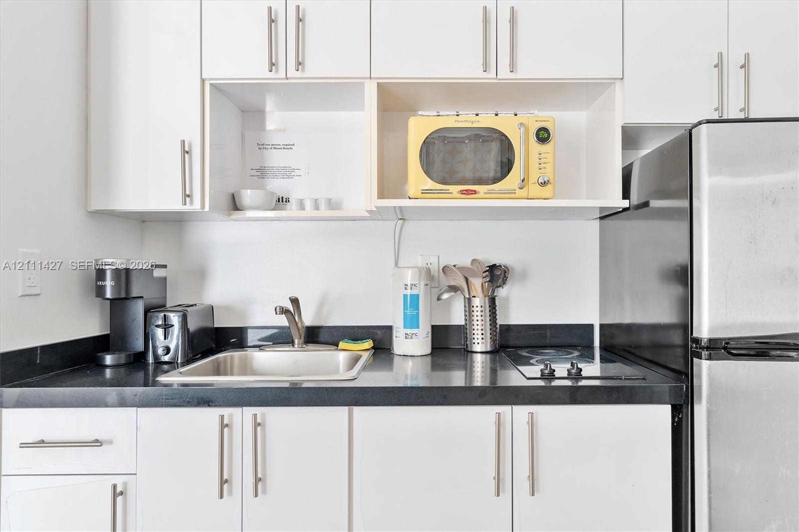 Kitchenette featuring white cabinetry with brushed-metal pulls, solid-surface dark countertops, a stainless steel undermount sink, and an integrated e
