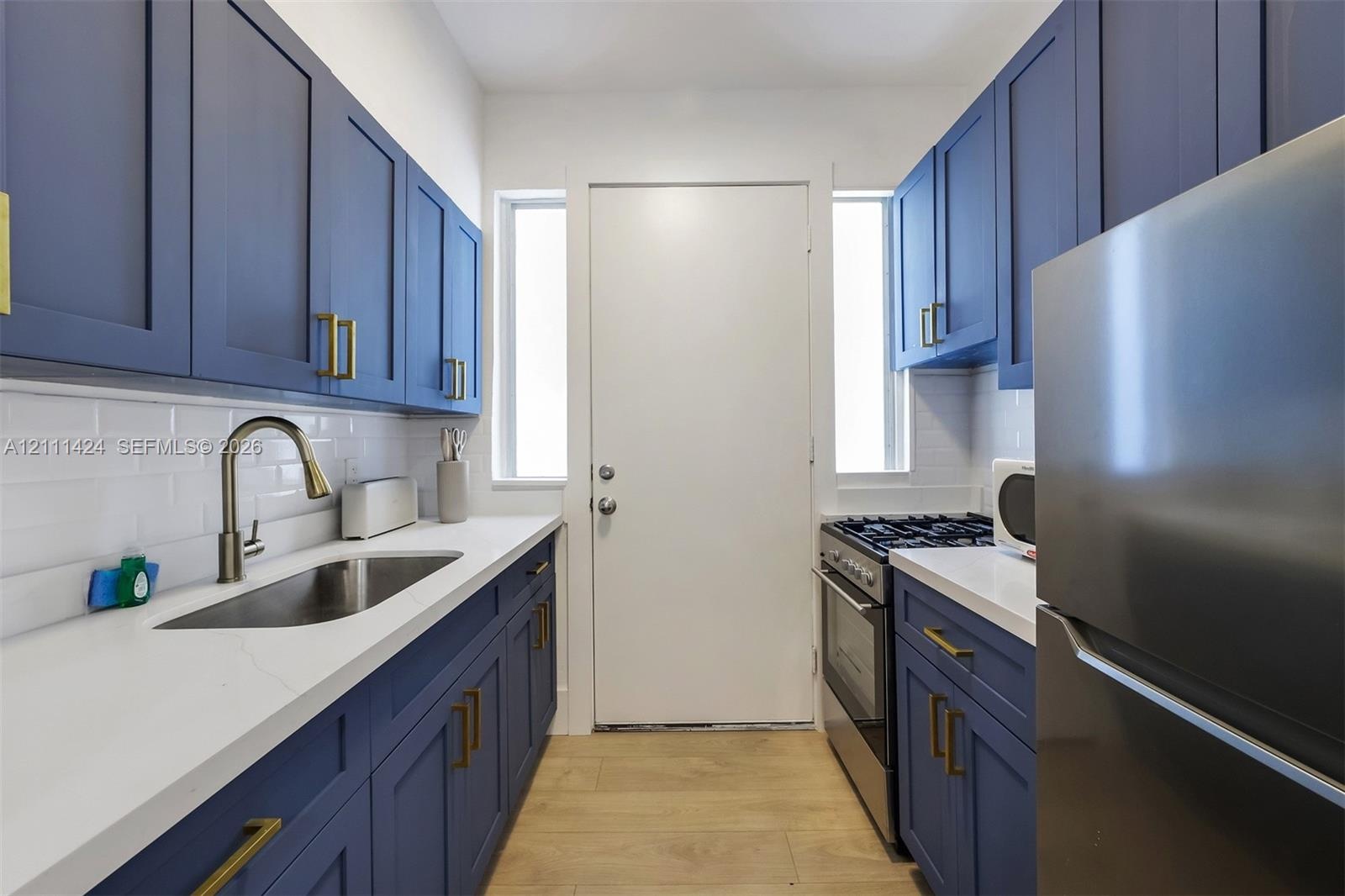 Galley kitchen featuring matte blue shaker-style cabinetry, gold-tone hardware, white subway tile backsplash, solid-surface countertops, and wood-fini