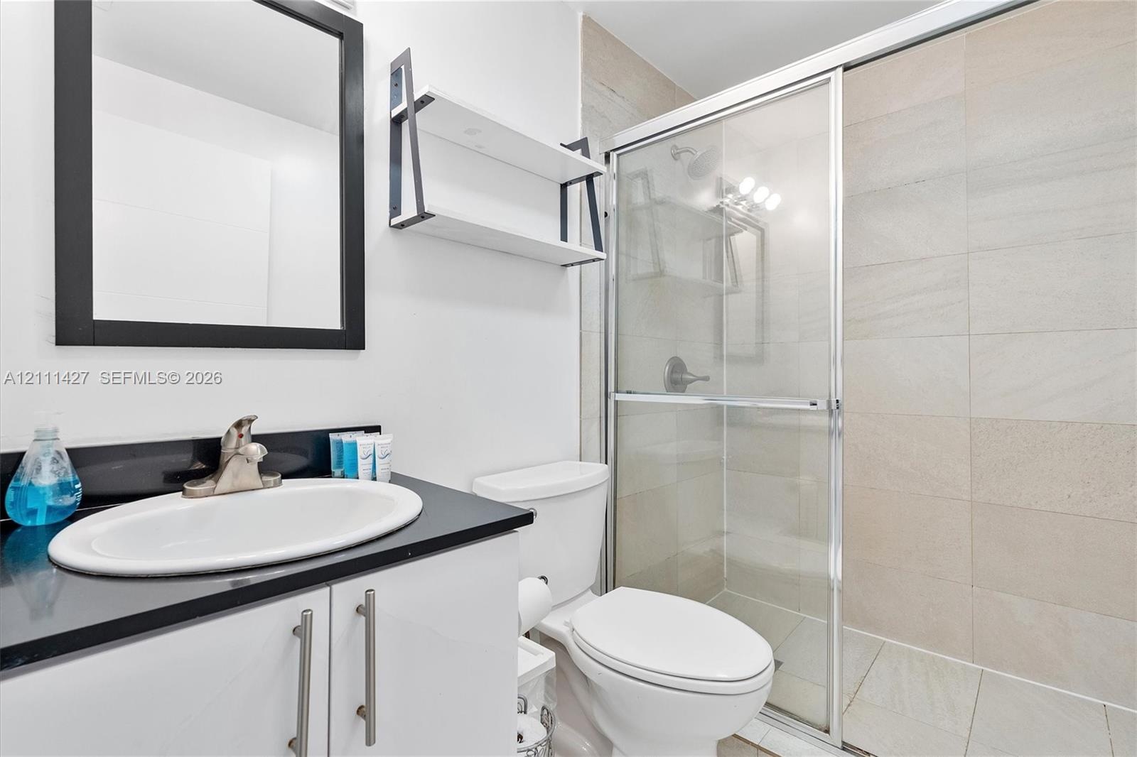 Bathroom featuring a vanity with a dark countertop and oval undermount sink, black-framed mirror, mounted shelving, and a shower enclosure with large-