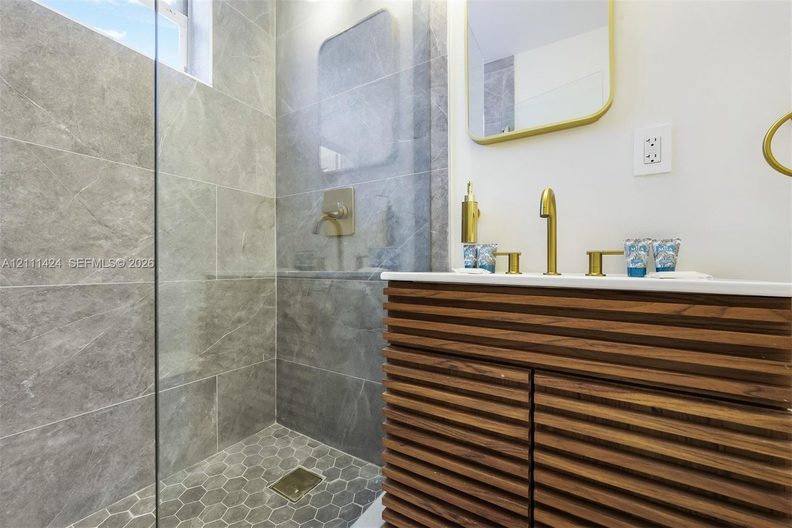 Floating vanity with horizontal wood-slat cabinetry, large-format grey stone-patterned wall tile, frameless glass shower enclosure, brass-finish hardw