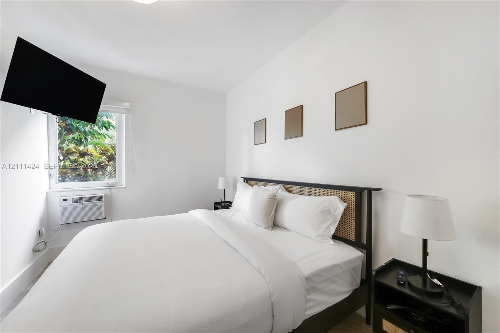 Bright bedroom featuring neutral walls, a single-pane window with exterior greenery, and an installed wall-mounted air conditioning unit