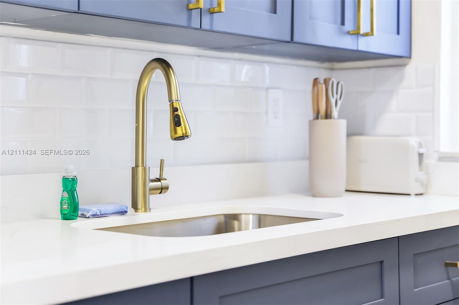 Blue shaker-style cabinetry with brass-finish hardware, undermount stainless steel sink, gold-tone gooseneck pull-down faucet, white solid-surface cou