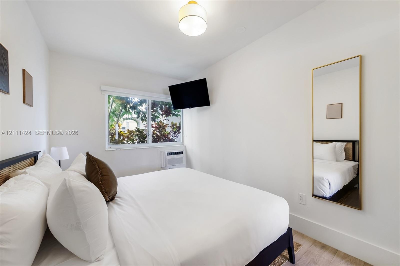 Bedroom featuring neutral walls, wood-finish flooring, a wall-mounted climate control unit, and a window with exterior greenery