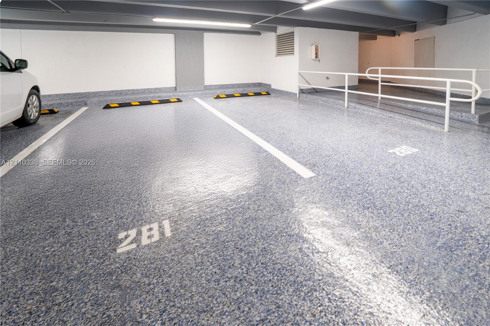 Covered parking garage featuring high-gloss speckled epoxy flooring, painted concrete walls, overhead strip lighting, and a white metal safety railing