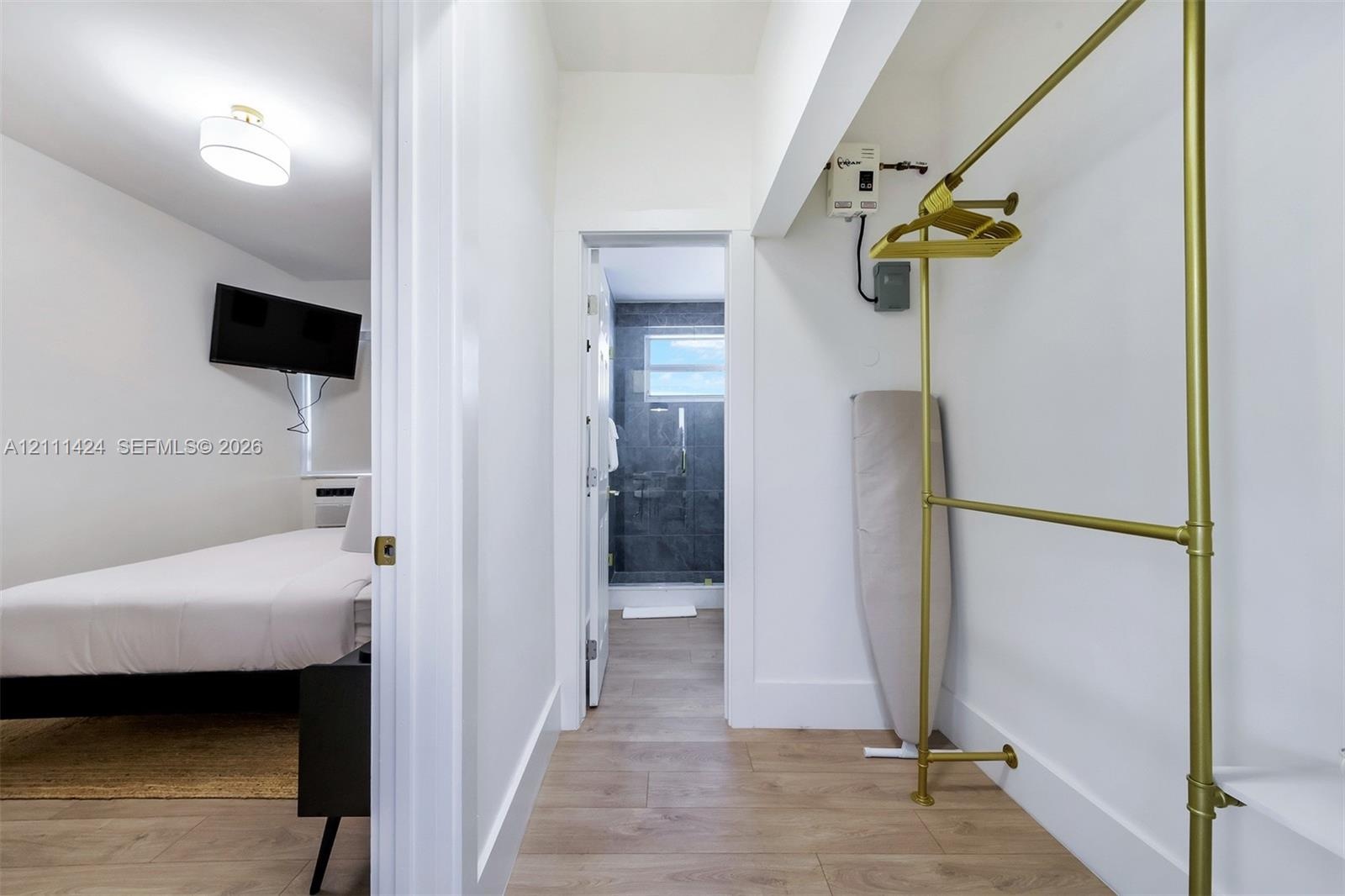 Hallway with wood-finish flooring leading to the bathroom, with a view into a bedroom and a gold-tone clothing rack