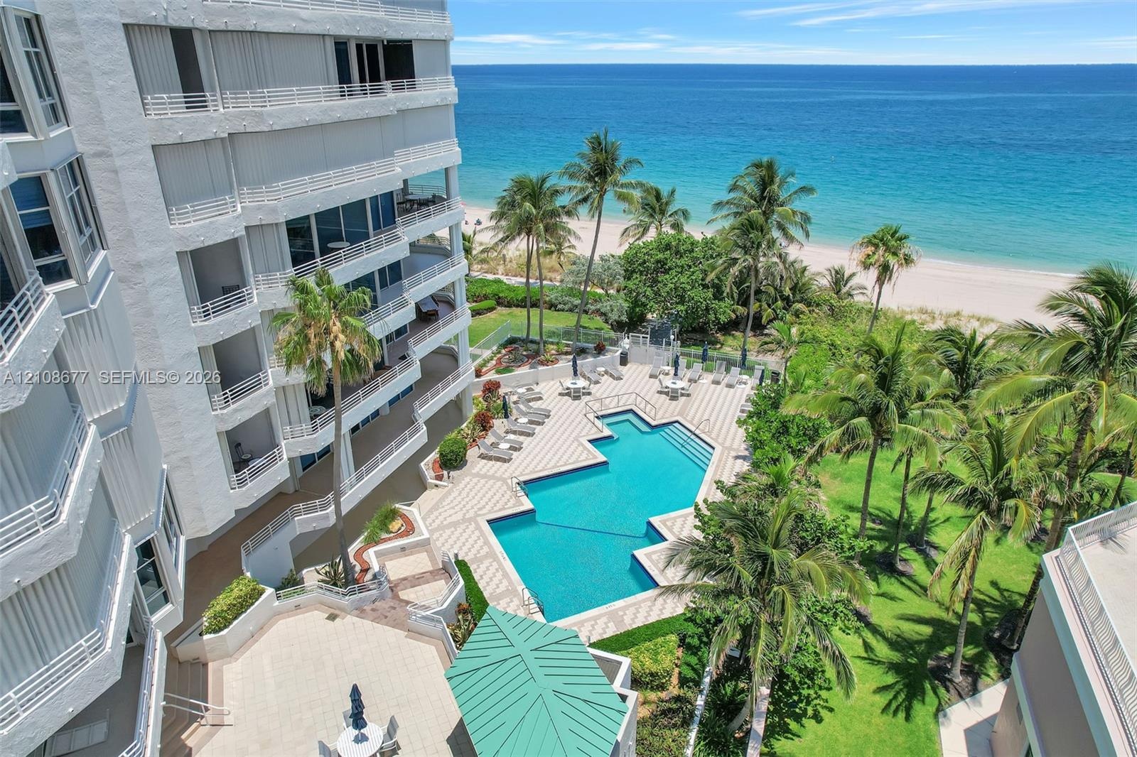 Multi-story residential building with private balconies overlooking an outdoor swimming pool, paved pool deck, manicured grounds with palm trees, and 