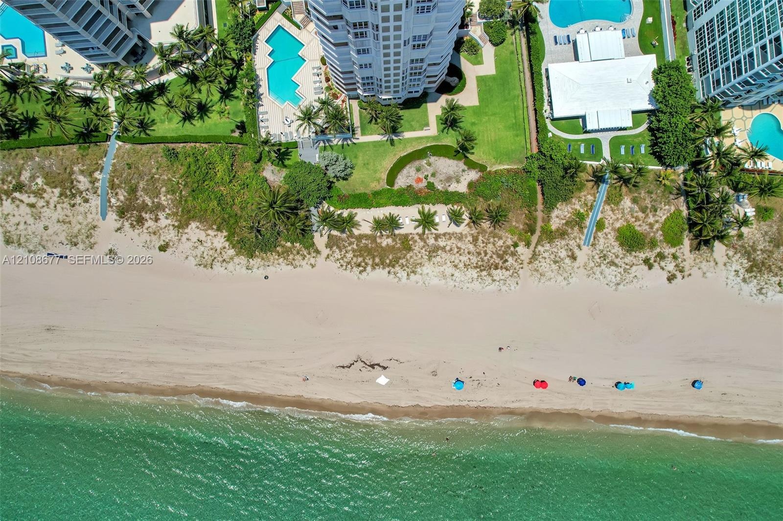 Aerial perspective of a coastal high-rise property featuring direct beach access, multiple outdoor swimming pools, manicured green space, and mature t