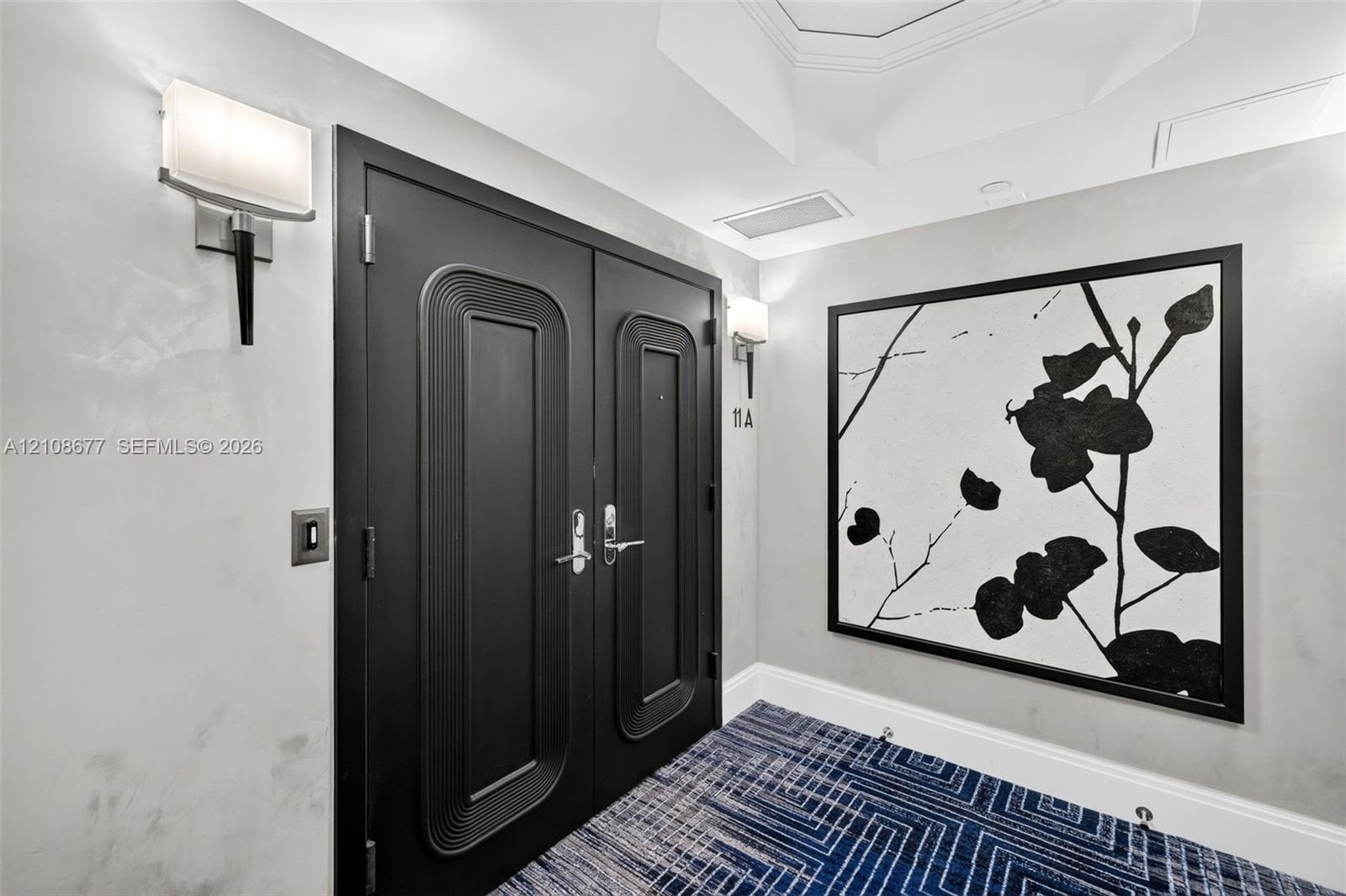 Double-door entryway featuring matte black hardware, geometric patterned carpet, dual wall-mounted light fixtures, and a coffered ceiling