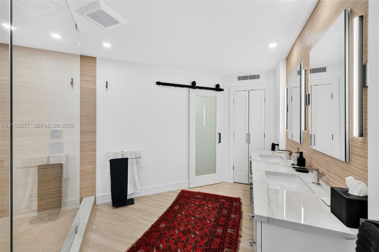 Double vanity with stone-surfaced countertops, frameless glass-enclosed shower, textured wood look wall tile, wall sconces, and frosted glass sliding 