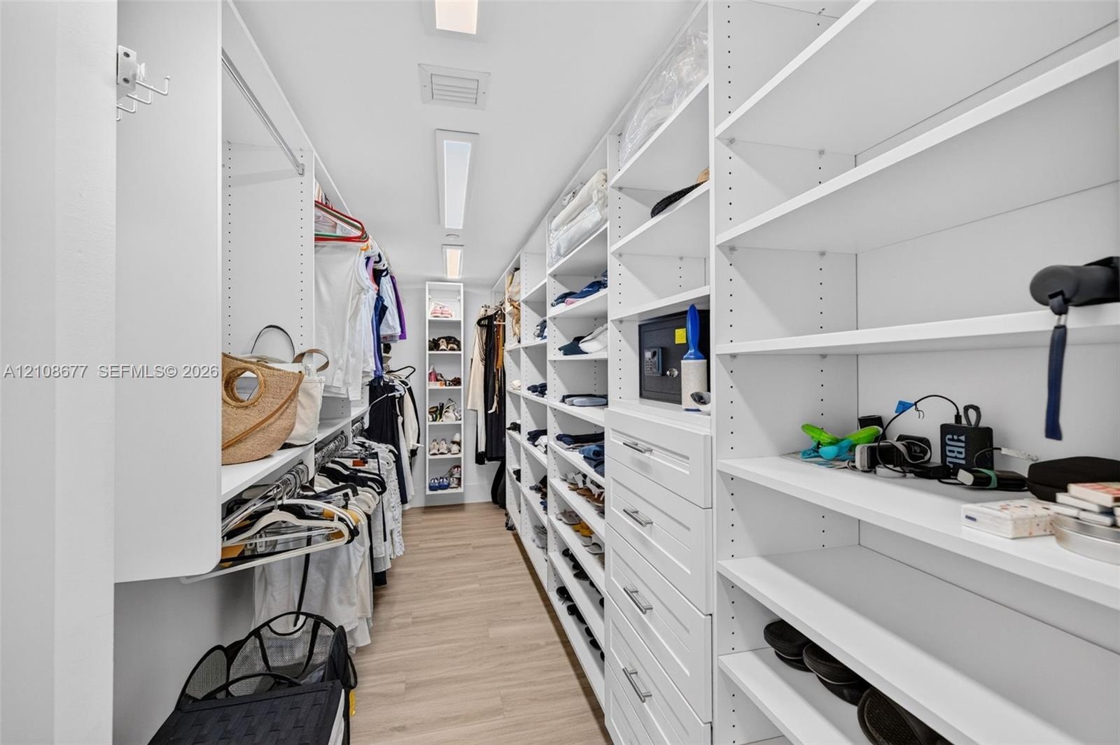 View toward long side of the custom walk-in closet featuring floor-to-ceiling white shelving, integrated cabinetry with drawers, multiple hanging rods