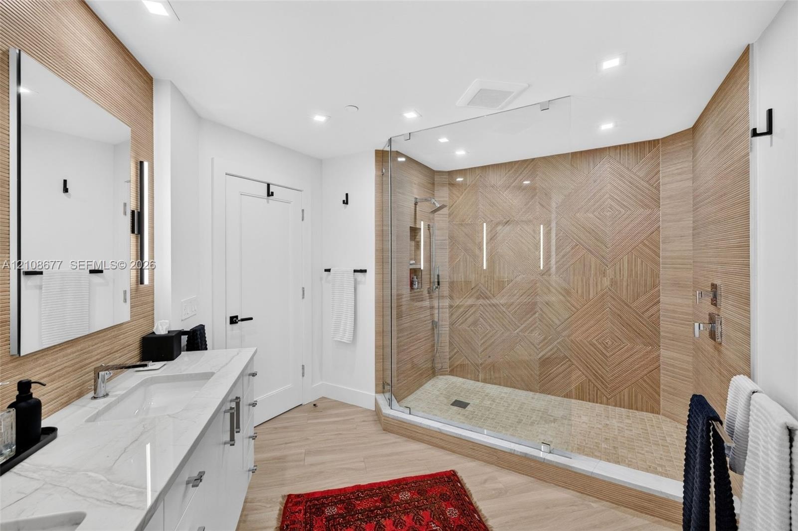 Modern bathroom featuring a dual-sink vanity with stone counters, a glass-enclosed shower with geometric tile work and built-in shelving, and wood-loo
