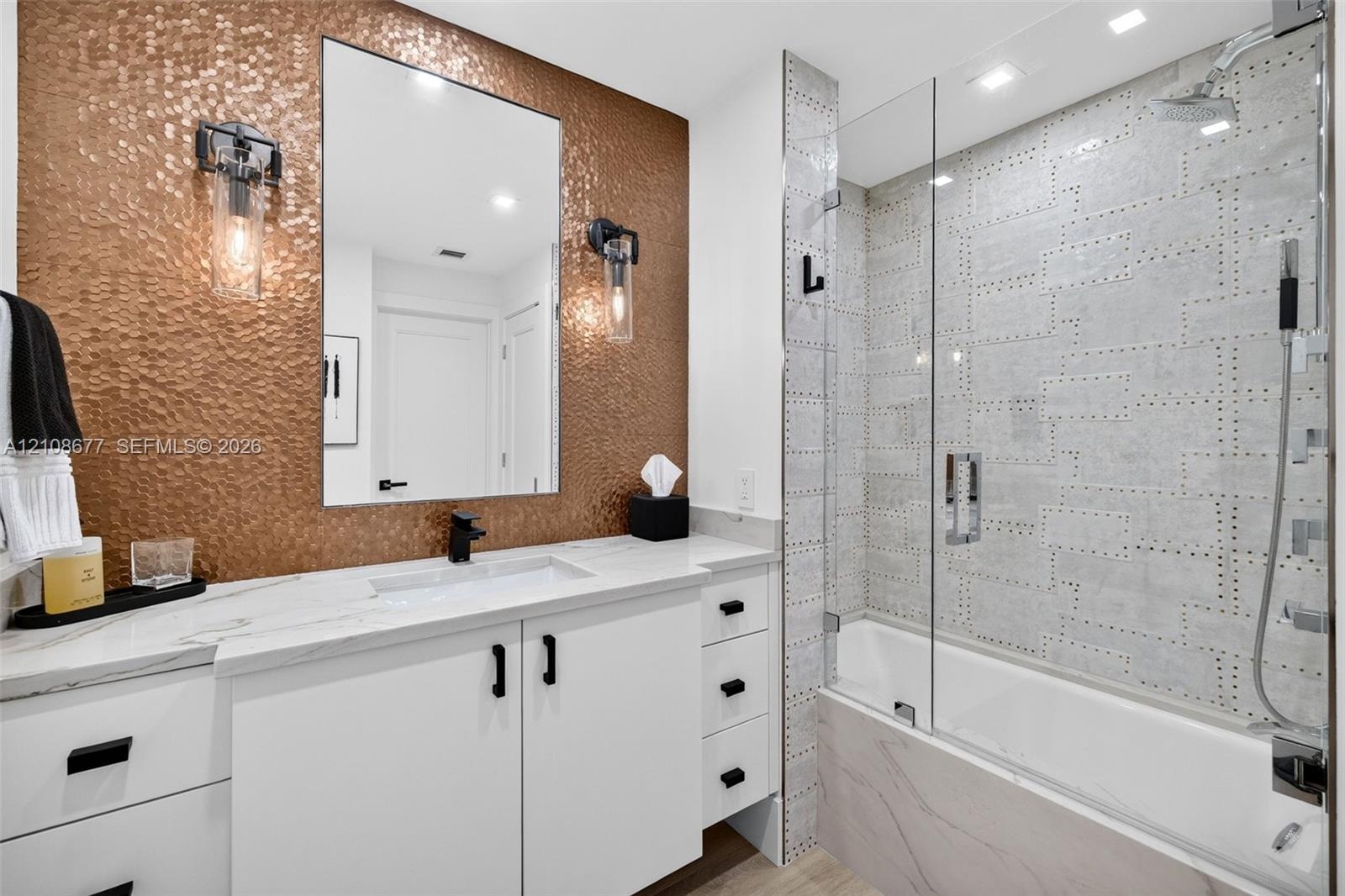 Bathroom featuring a hammered metallic tile accent wall, white floating vanity with stone counters, undermount sink, frameless glass shower enclosure,