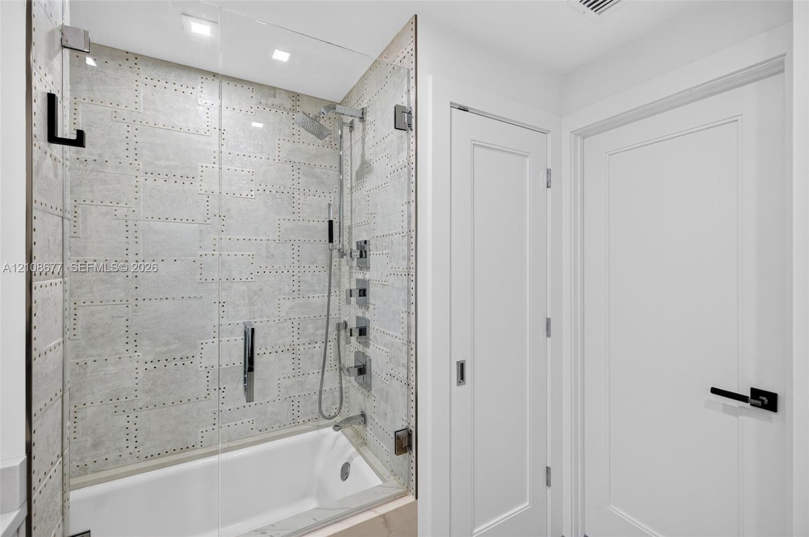 Shower enclosure featuring luxury patterned wall tile, frameless glass door, adjustable-height handheld showerhead, wall-mounted faucet controls, and 
