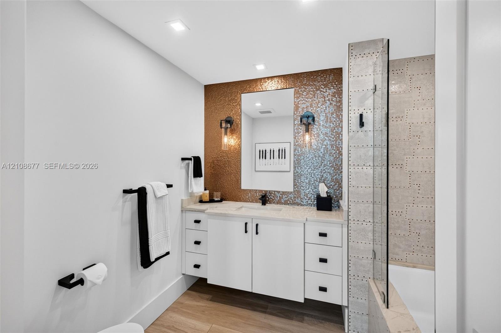 Bathroom w/ custom wall hung white vanity with matte black hardware, metallic mosaic tile accent wall, recessed lighting, and a glass-enclosed shower 