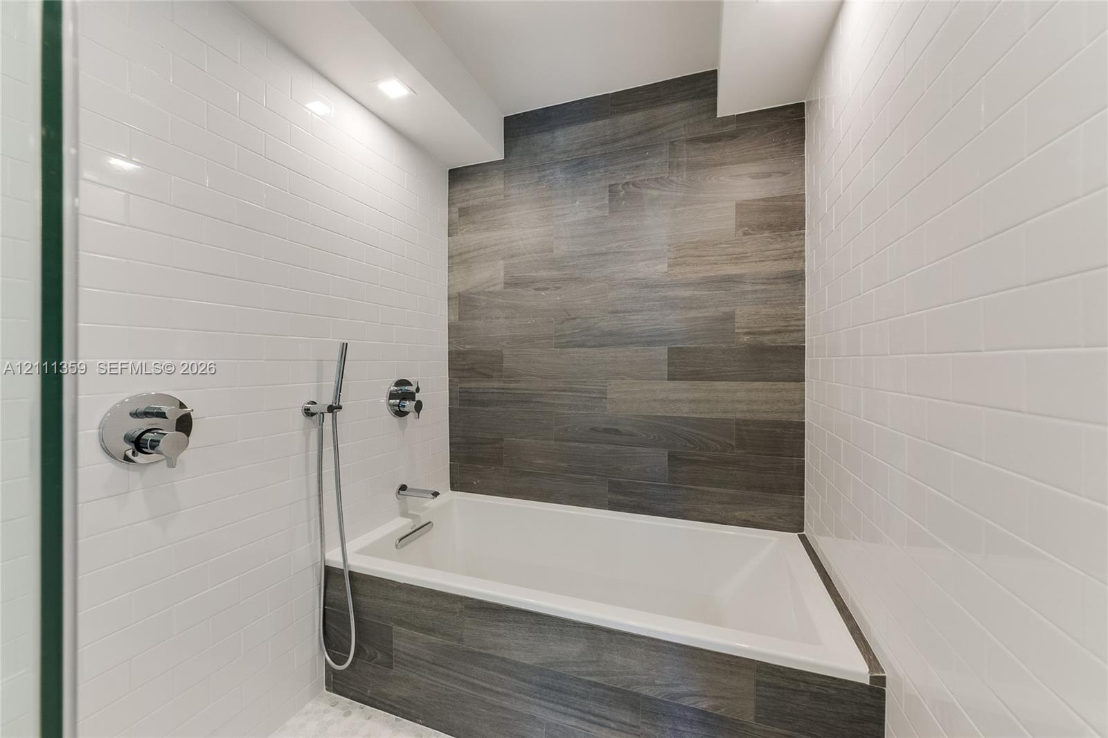 Built-in deep soaking tub featuring wood-finish tile surround, white subway tile walls, chrome shower fixtures, and recessed ceiling lighting