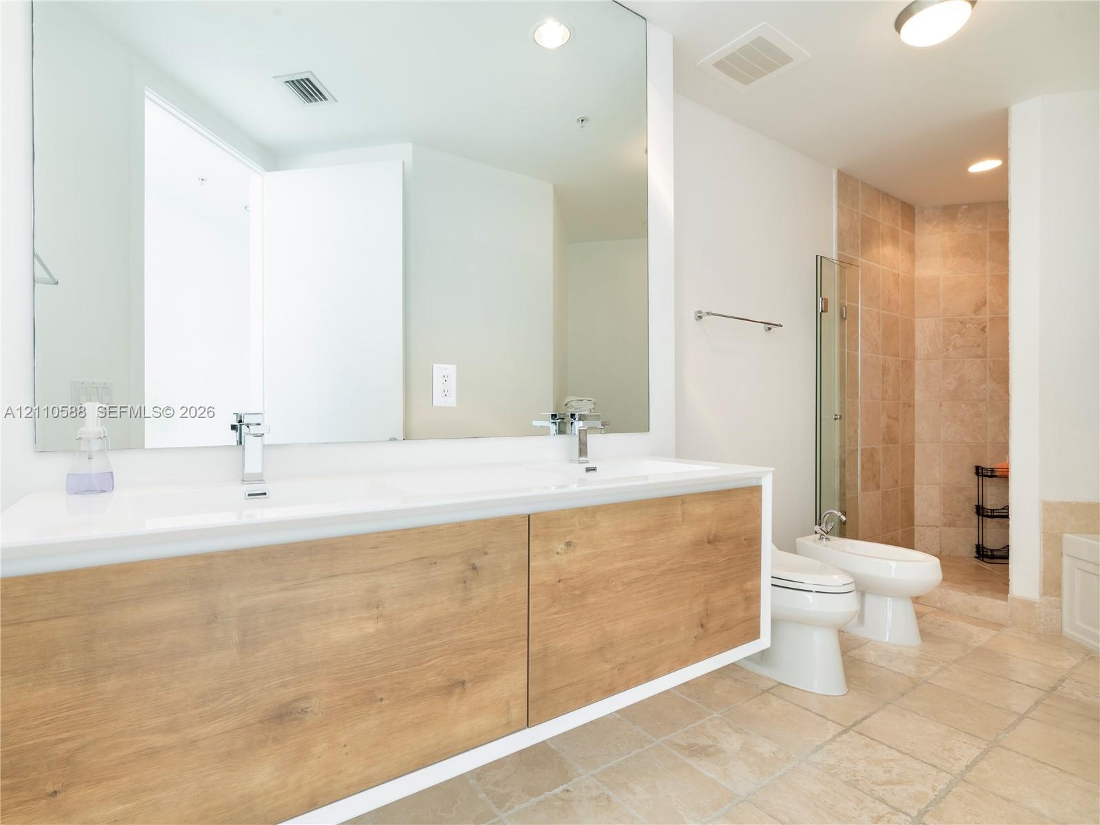 Floating dual-sink vanity with wood-finish cabinetry, expansive wall-mounted mirror, stone-tile flooring, and glass-enclosed shower