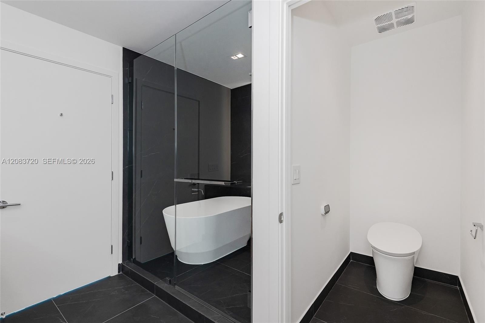 Freestanding soaking tub enclosed in a glass shower stall with dark tile surround, complemented by dark floor tiling and a white high-profile toilet