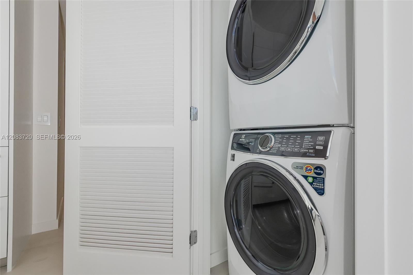 Stacked white front-loading laundry appliances enclosed within a closet featuring a white louvered door