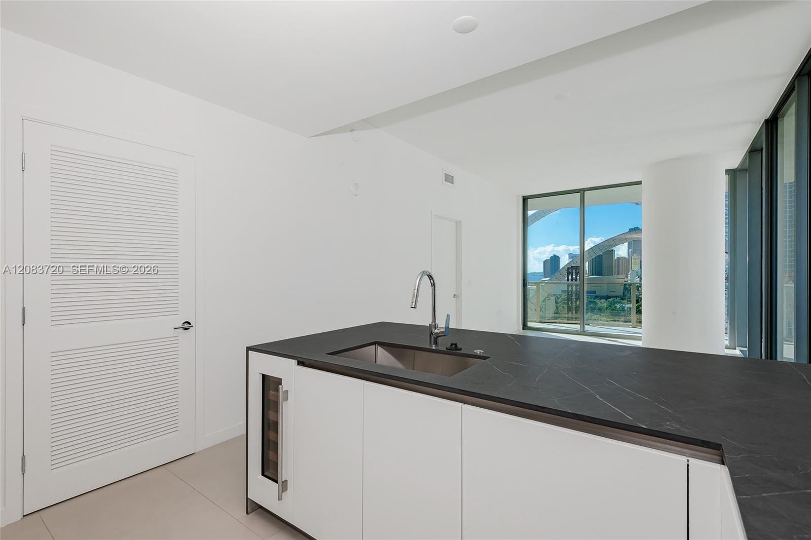 Kitchen island featuring a dark stone countertop, integrated wine refrigerator, stainless steel undermount sink, and high-arc faucet, with white louve