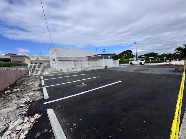 Asphalt-paved parking area featuring marked spaces, concrete parking bumpers, a perimeter metal fence, and an exterior structure with a covered entran