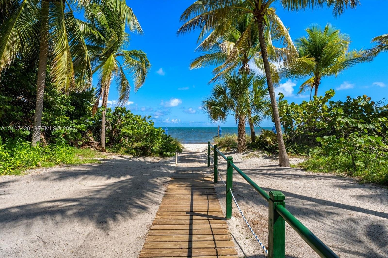 Sandy path featuring a wooden boardwalk, metal railings with rope accents, mature palm trees, and coastal water access