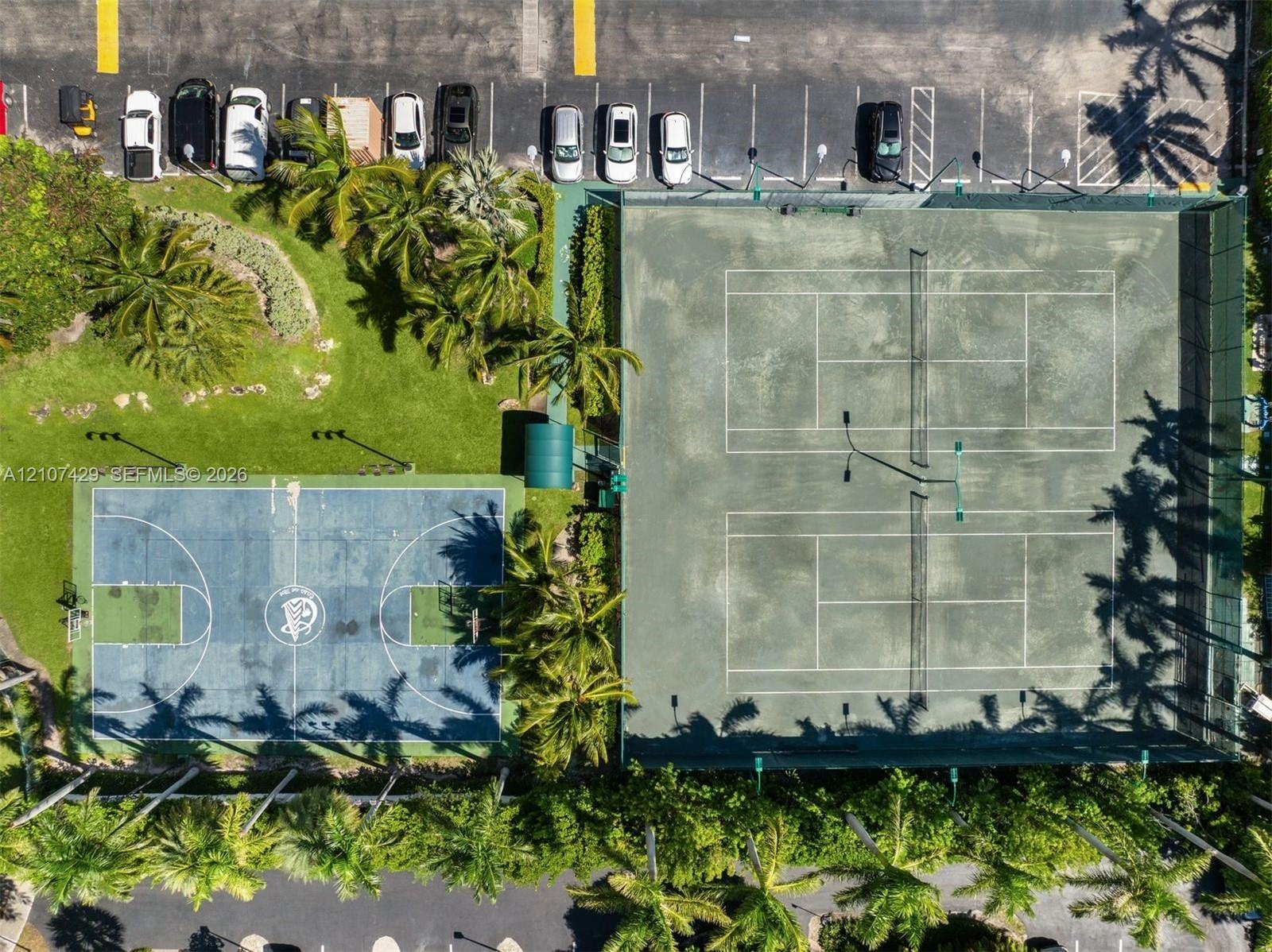 Outdoor recreational complex featuring two fenced tennis courts and a hard-surface basketball court surrounded by mature palm trees and landscaped gre