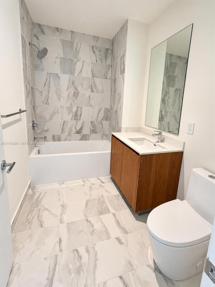 Bathroom featuring large-format tile flooring, wood-finish vanity with a rectangular sink, wall-mounted mirror, and a tile-surround soaking tub with a