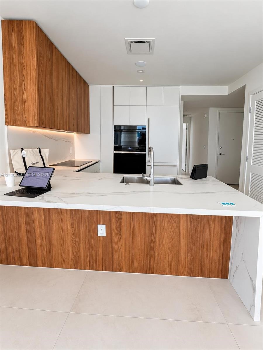 Modern kitchen featuring a waterfall-edge stone countertop, wood-finish cabinetry, built-in wall oven, undermount sink with a gooseneck faucet, and la