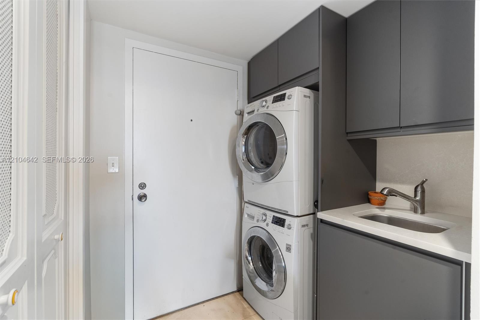 Laundry area featuring charcoal-toned cabinetry, light-toned stone-finish countertop, integrated sink with a gooseneck faucet, and stacked appliance u
