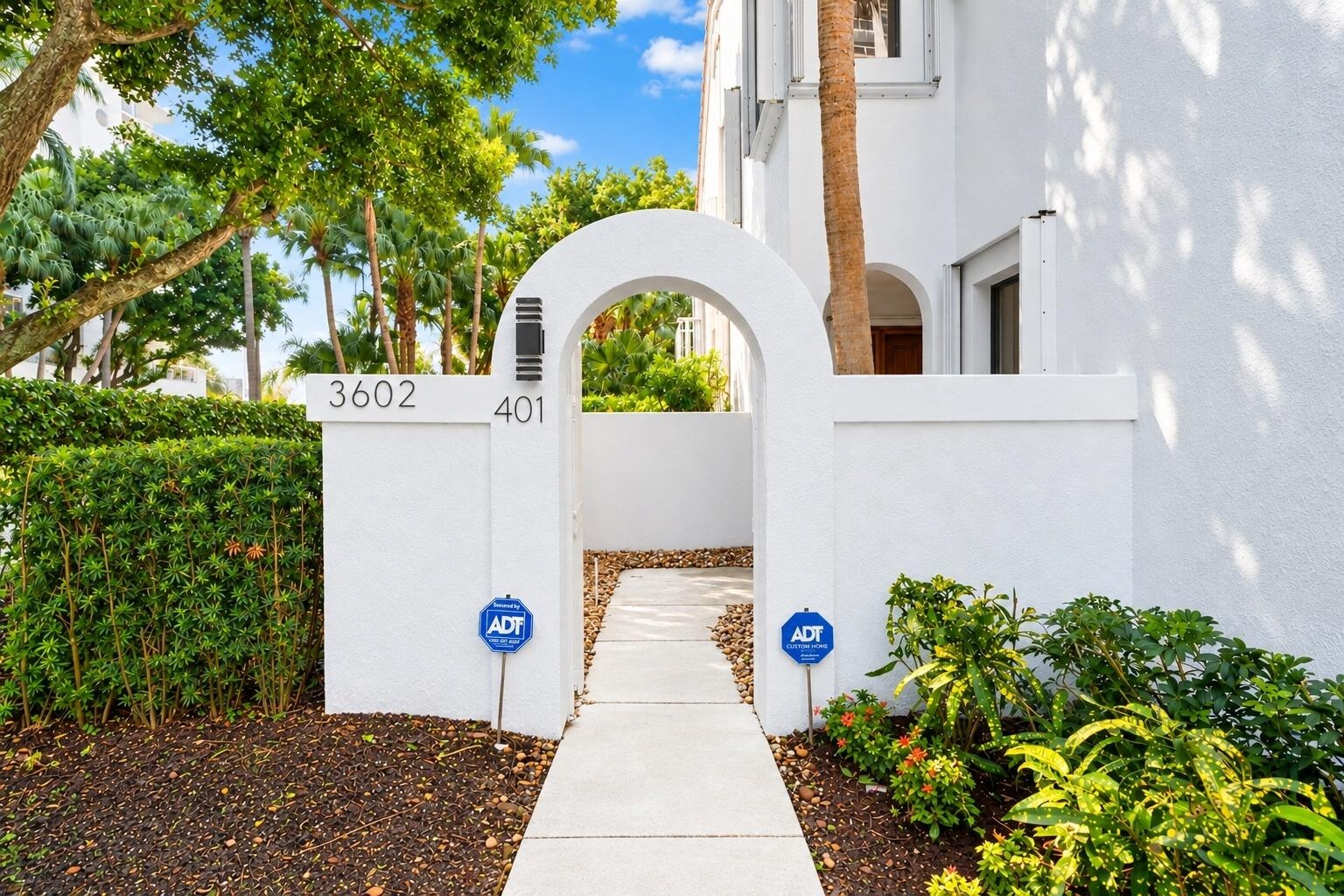 Stucco-finished exterior wall featuring an arched entryway, concrete walkway, wall-mounted exterior lighting, and landscaped perimeter hedges
