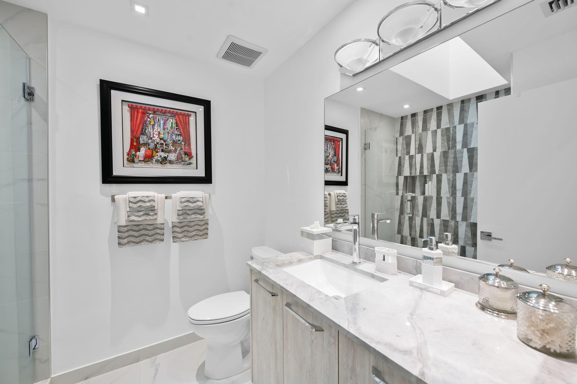 Vanity featuring a light-toned wood-finish cabinet and stone-surface countertop with an integrated basin, large wall-mounted mirror, geometric tile sh